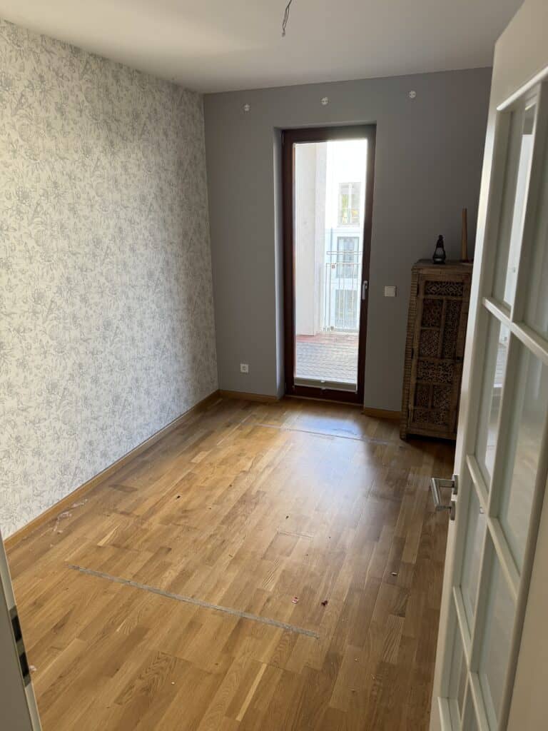 Leeres Zimmer mit hellem Holzboden, gemusterter Tapete an einer Wand, einem einzelnen Holzschrank, einer Glastür und einem Balkon, der durch ein hohes Fenster zu sehen ist.