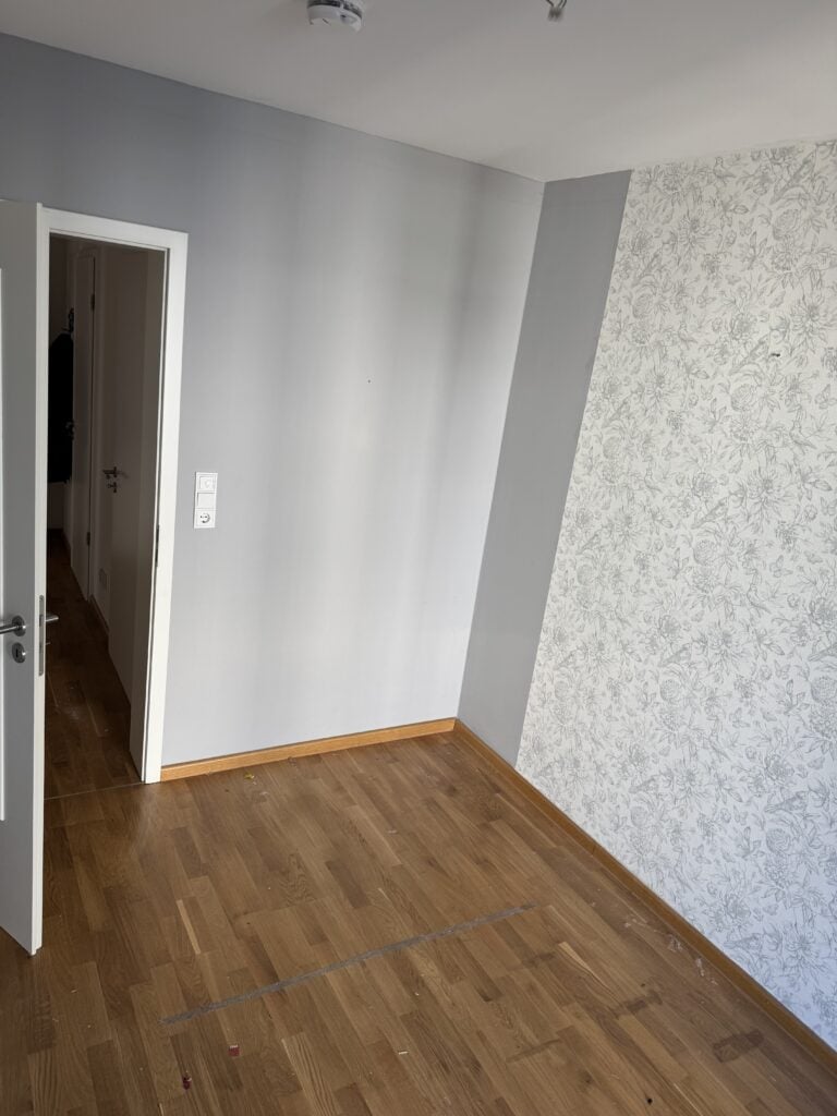 Leerer Raum mit hellgrauen Wänden, einer Akzentwand mit Blumenmuster, Holzboden und einer offenen weißen Tür, die zu einem Flur führt.