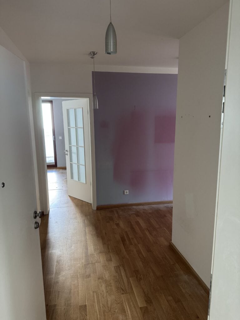 Ein Flur mit Holzfußboden, weißen Wänden, einer Hängelampe und einer offenen Tür, die zu einem Raum mit einer teilweise gestrichenen lila Akzentwand führt.