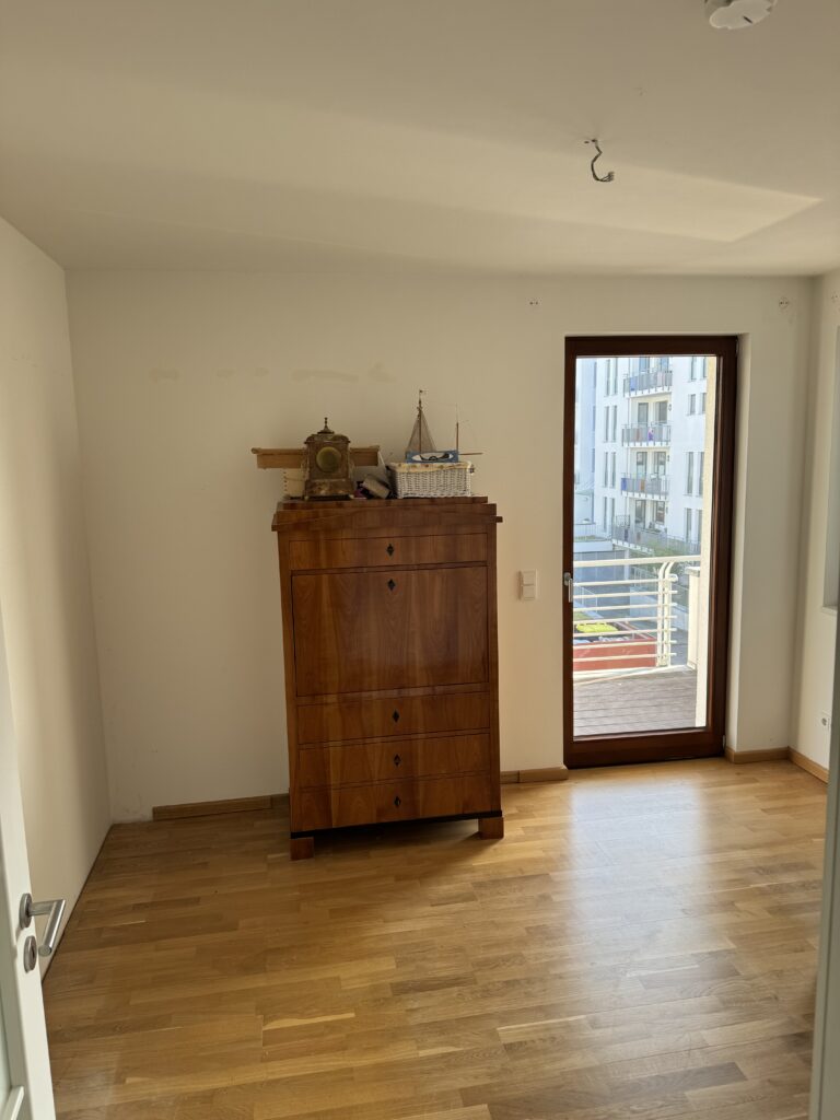 Ein kleiner, weitgehend leerer Raum mit Holzboden, einer Holzkommode an der Wand und einer Glastür, die auf einen Balkon führt.