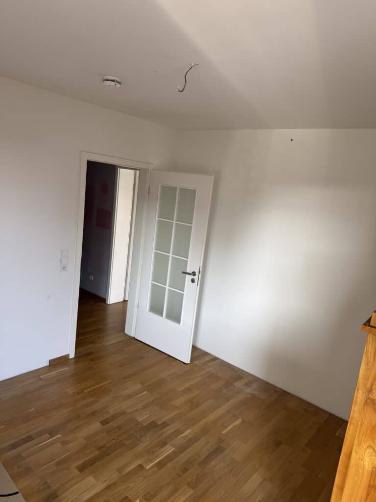 Leerer Raum mit weißen Wänden, Holzboden, einer offenen Tür mit Glasscheiben und einem herausragenden Deckendraht; natürliches Licht fällt von links ein.