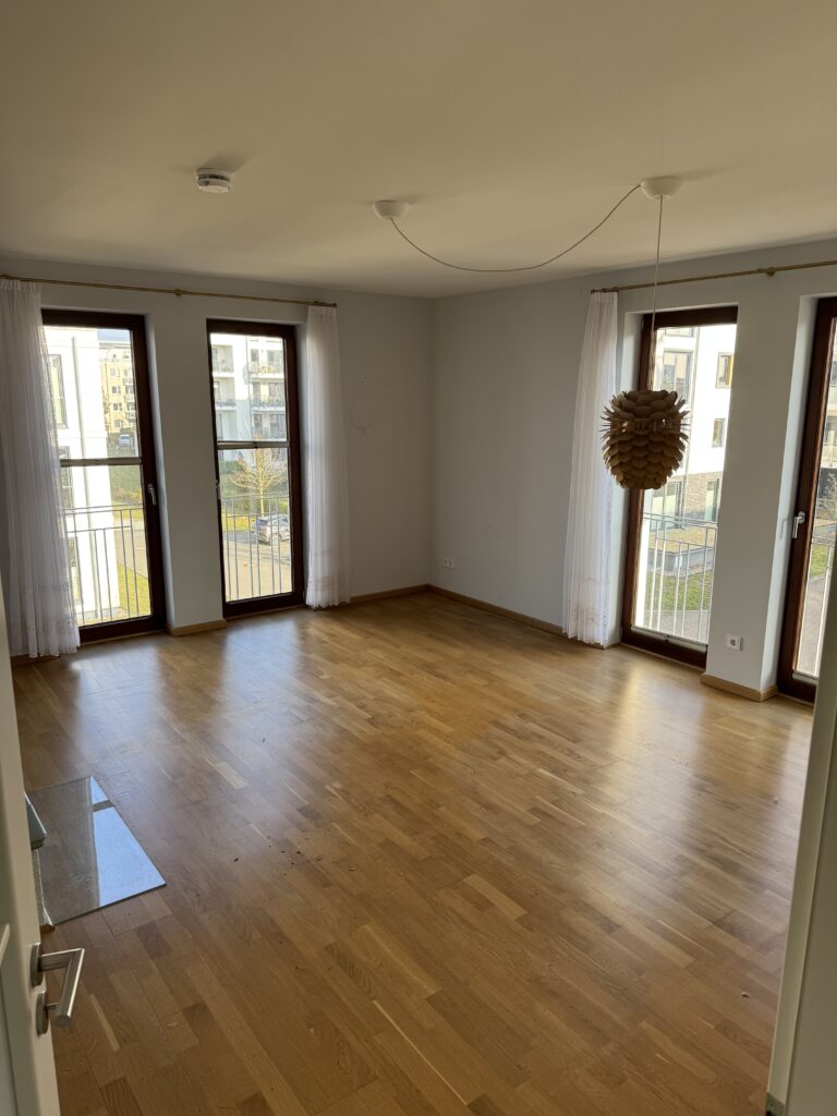 Leeres Zimmer mit Holzboden, drei großen Fenstern mit weißen Vorhängen und einer Deckenleuchte. Durch die Fenster ist ein Balkon zu sehen.