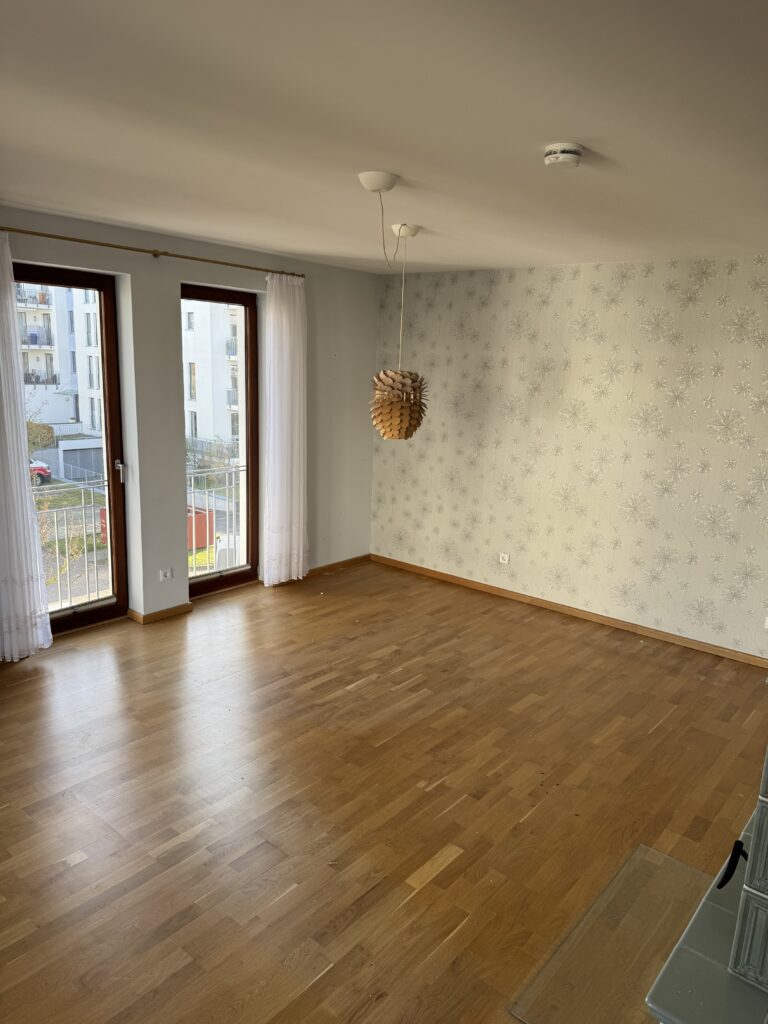 Leeres Zimmer mit hellem Holzboden, großem Fenster und Tür mit weißen Vorhängen, Blumentapete an einer Wand und einer Deckenleuchte.