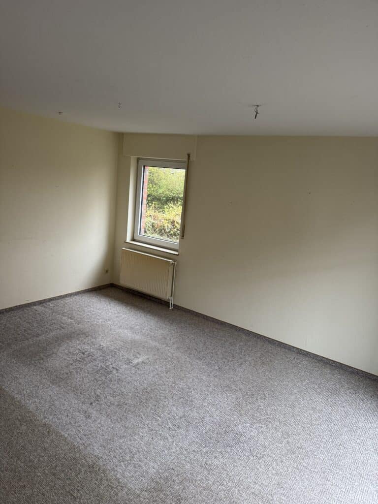 Leeres Zimmer mit beigen Wänden, einem einzigen Fenster mit einem Heizkörper darunter und einem grauen Teppichboden, der einige Flecken und Abnutzungserscheinungen aufweist.