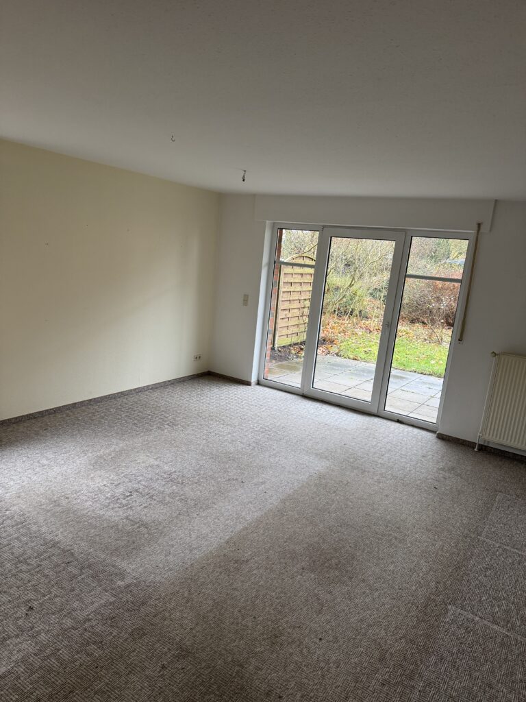 Leeres Zimmer mit Teppichboden und großen Glastüren, die zu einer Terrasse und einem Gartenbereich führen; neutrale Wände und ein Heizkörper auf der rechten Seite.