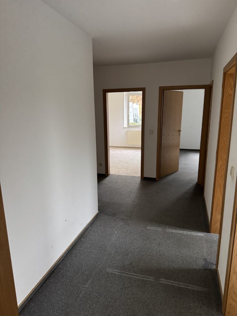 Ein Flur mit grauem Teppichboden führt zu zwei offenen Holztüren, die den Blick auf Zimmer mit Teppichboden und ein Fenster mit einem Heizkörper im Hintergrund freigeben.