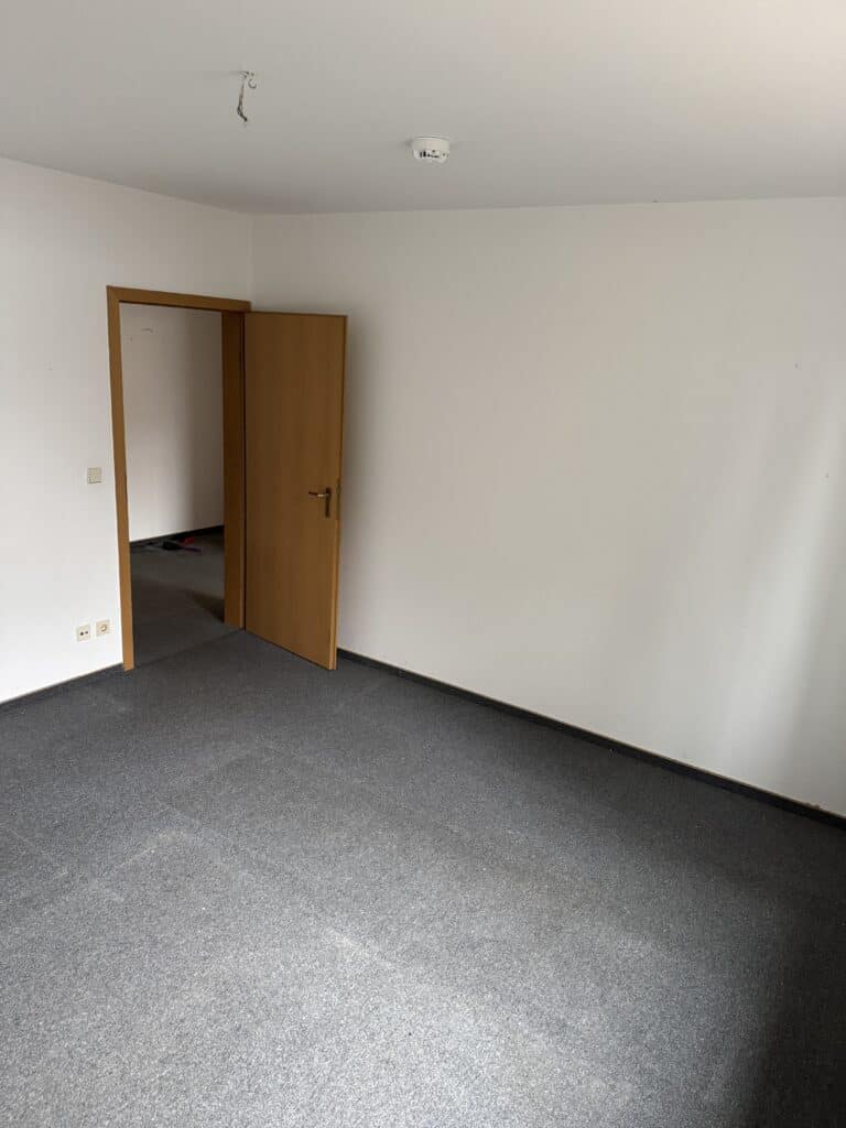 Ein leerer Raum mit grauem Teppich, weißen Wänden, einer teilweise geöffneten Holztür und einem Rauchmelder an der Decke.