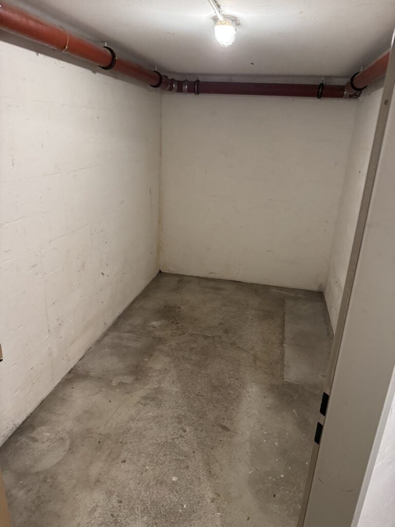 Kleiner, leerer Betonraum mit kahlen Wänden, freiliegenden Rohren an der Decke, einer einzigen Glühbirne und einem rauen Betonboden.