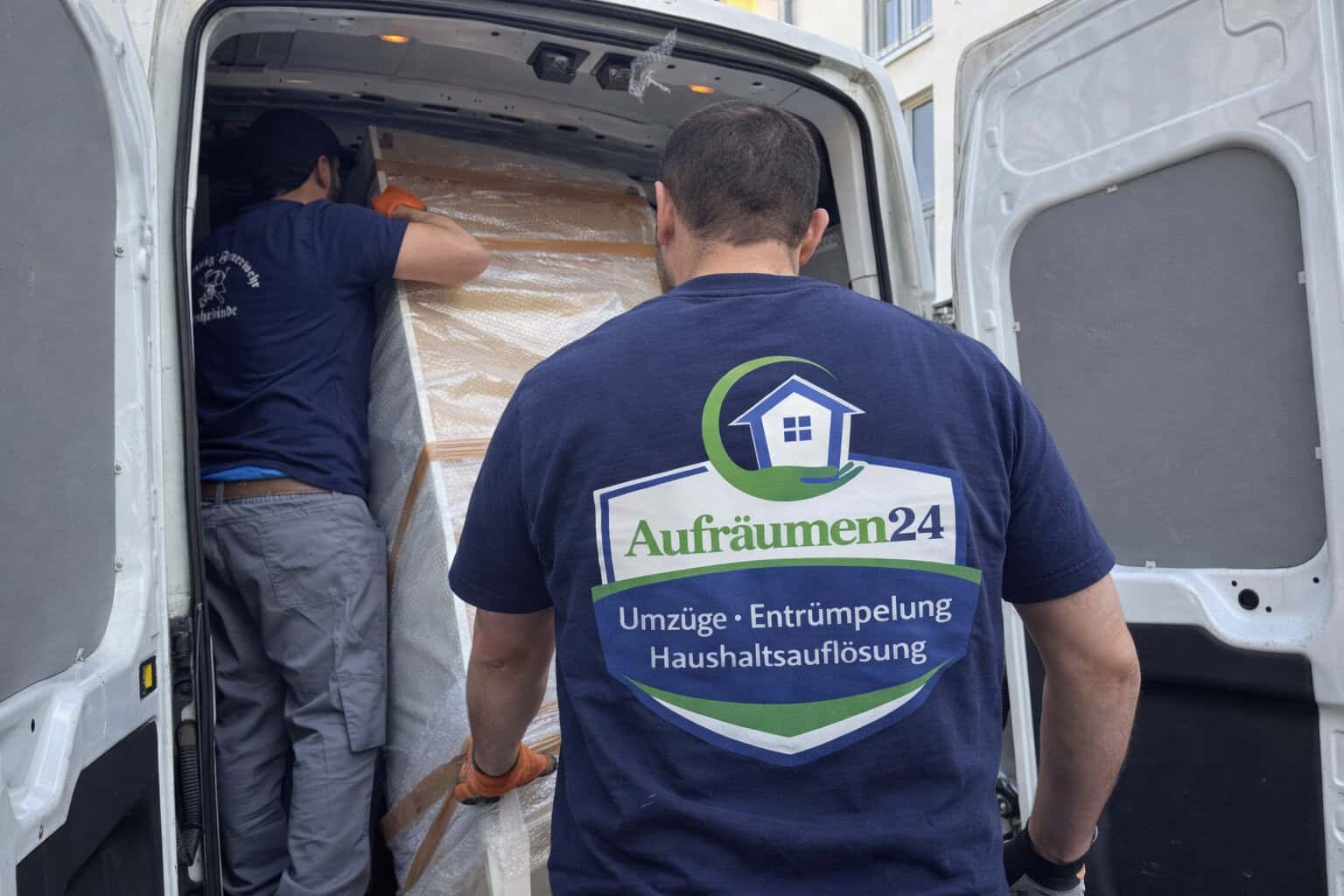 Zwei Umzugshelfer in Aufräumen24-Hemden laden vorsichtig einen großen, verpackten Gegenstand in einen weißen Transporter - ideal für Privatumzüge oder Möbeltransporte. Die Türen des Lieferwagens sind offen, eine weitere Person im Inneren hilft mit.